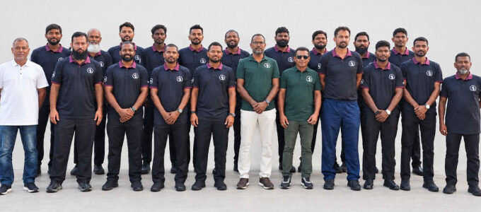 The Maldives national cricket squad and team management lined up together outdoors against a plain white wall, most wearing dark navy Cricket Board of Maldives polo shirts, ahead of departure for a training camp in Sri Lanka.