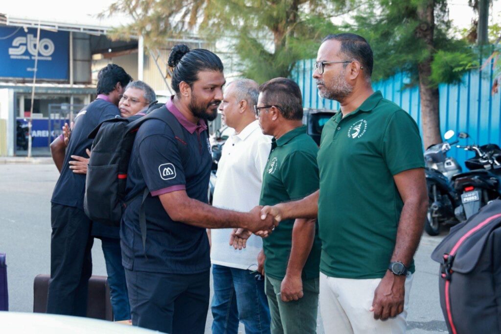 A Cricket Board of Maldives squad member in a navy training polo shakes hands with an official in a green CBM polo during a send-off at the departure point, with other officials standing nearby and a second squad member embracing someone in farewell; a street scene with motorbikes and shopfronts is visible behind them.