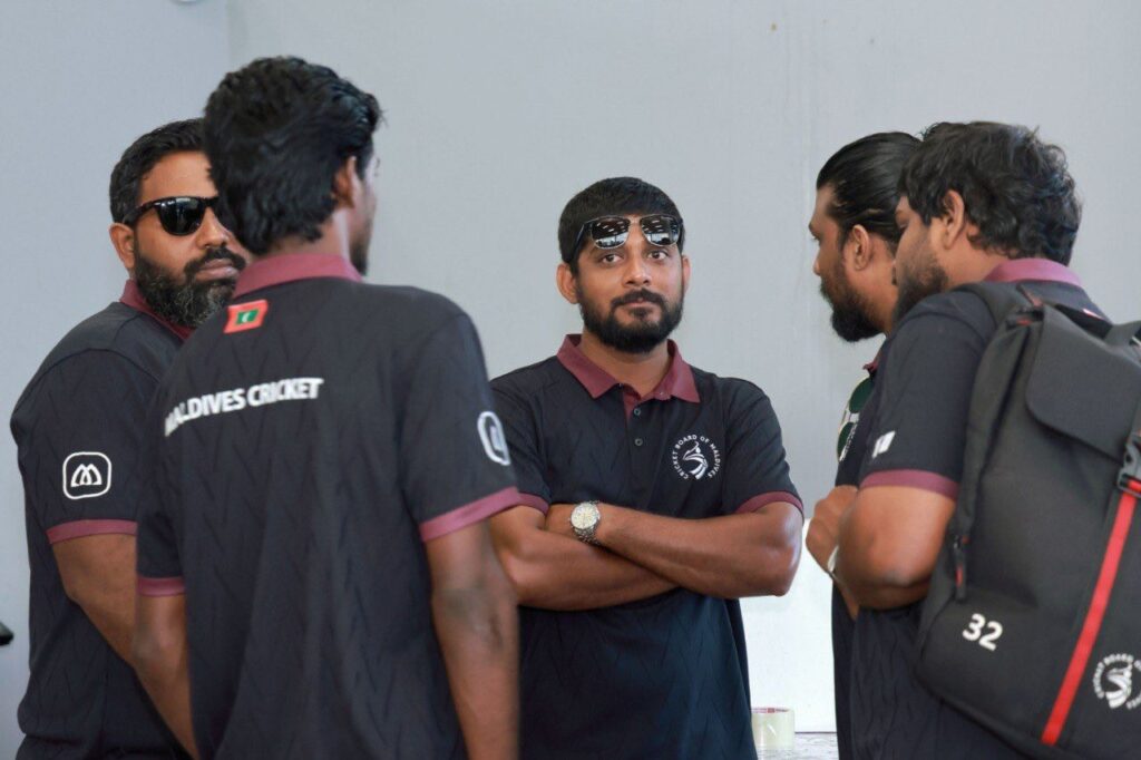 Five or six squad members in dark Cricket Board of Maldives training polos with maroon collars stand together indoors against a plain wall, several carrying backpacks; one faces the camera with sunglasses pushed up on his head, and the Maldives Cricket wordmark and flag are visible on the back of another player's shirt.