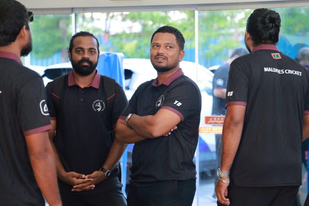 Two men in dark Cricket Board of Maldives polos with maroon collars talk indoors beside glass doors, with daylight and parked vehicles visible outside; two squad members are partly in frame, one with his back to the camera showing the Maldives Cricket wordmark.